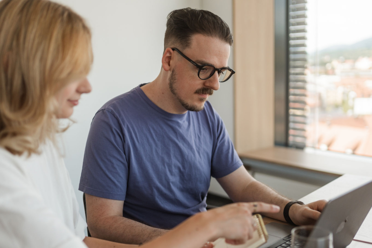 Ein Mann mit dunklen Haaren, Brille und Lila Shirt sitz mit einer blonden Frau, mit einer weissen Bluse, vor dem Laptop. Sie schauen beide in den Rechner.